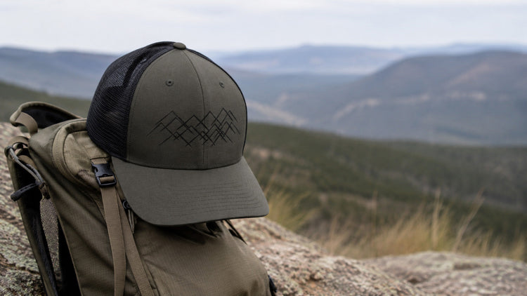 Casquette trucker kaki et noire Sortie Longue avec motif montagne, posée sur un sac de randonnée en pleine nature, paysage outdoor et reliefs en arrière-plan.