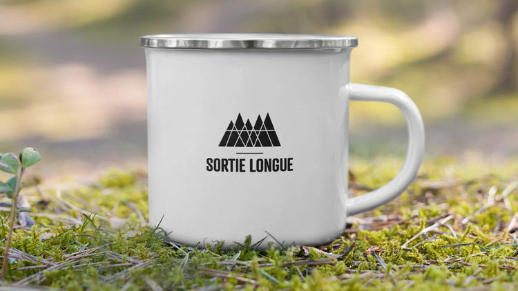 Mug métal émaillé blanc Sortie Longue avec logo montagne, posé dans la mousse en forêt, accessoire outdoor pour randonnée, trail et moments dehors.