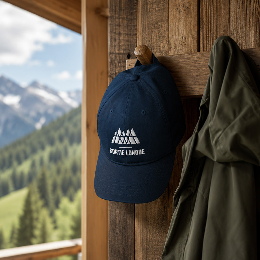 Casquette coton bio bleu marine Sortie Longue suspendue dans une entrée bois avec veste outdoor, ambiance montagne calme et naturelle