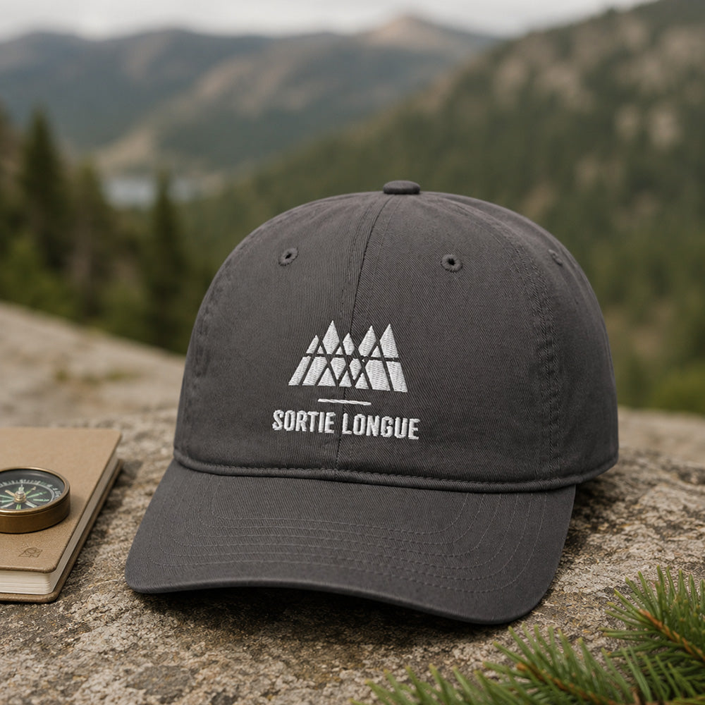 Casquette coton bio gris charbon Sortie Longue posée sur un rocher avec carnet, boussole et branche de sapin, ambiance randonnée montagne.