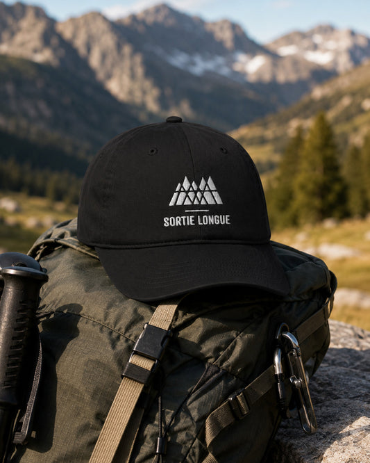 Casquette coton bio noire Sortie Longue posée sur un sac de randonnée, logo montagne brodé blanc, ambiance outdoor en montagne