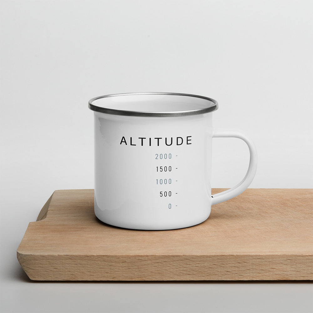 Mug émaillé blanc Sortie Longue avec design Altitude, échelle graphique minimaliste et graduations de hauteur, posé sur une planche en bois clair.