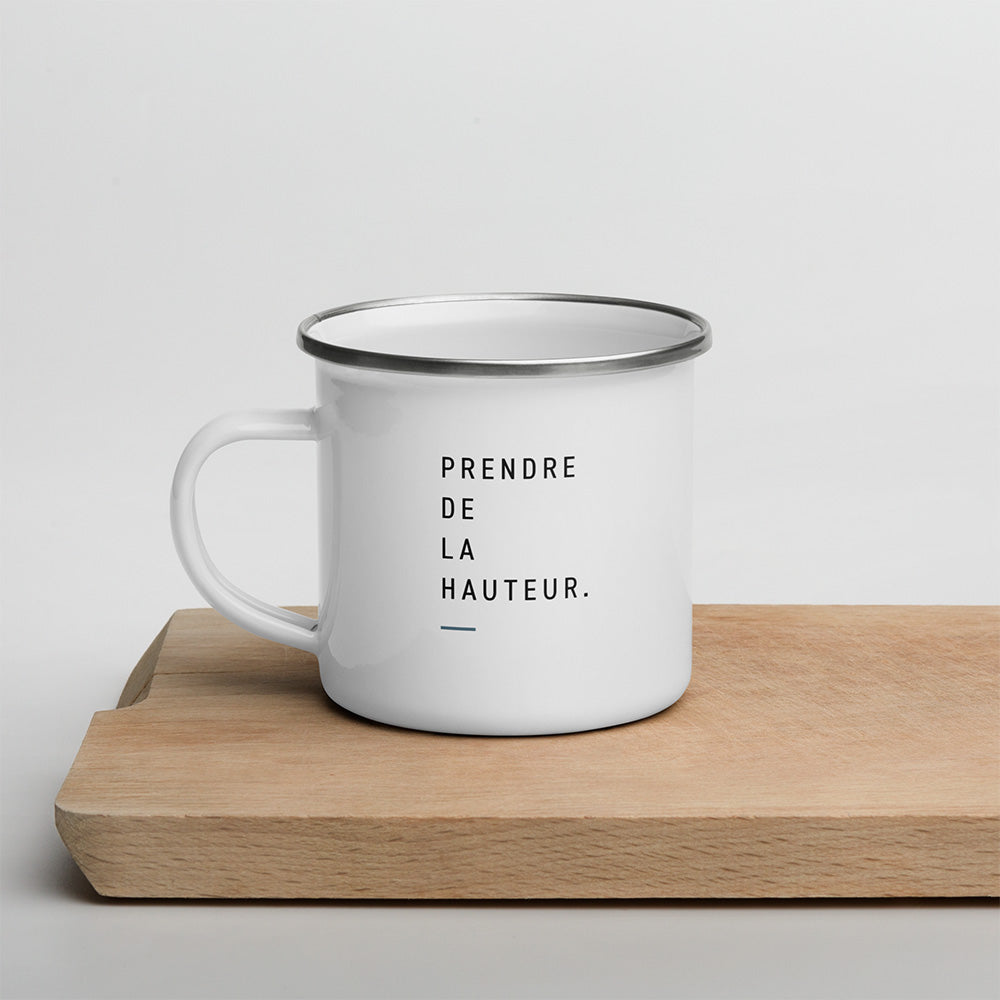 Mug émaillé blanc Altitude posé sur bois, avec la phrase Prendre de la hauteur imprimée en typographie sobre.