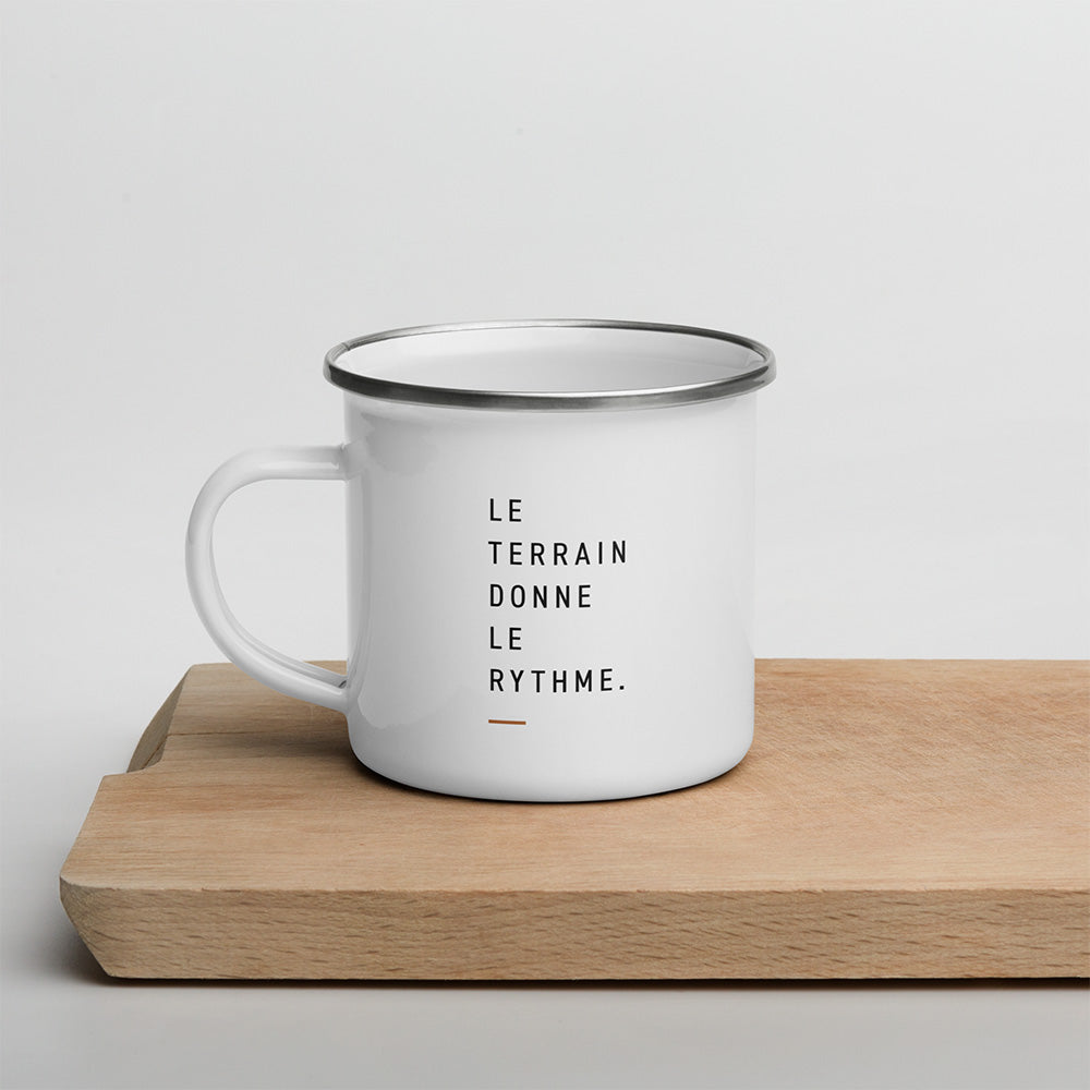 Mug émaillé blanc posé sur une planche en bois avec la phrase Le terrain donne le rythme, design minimaliste outdoor Sortie Longue.