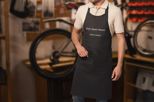Homme portant un tablier noir “Ride. Repair. Repeat.” dans un atelier vélo, en train de travailler sur son matériel.