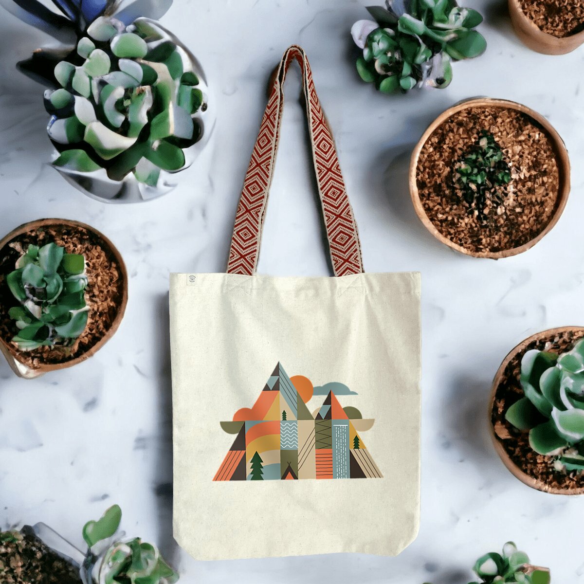 Tote bag coton recyclé Sortie Longue posé en extérieur, design géométrique montagne représentant sommets, route en lacets, chute d’eau et éléments naturels, usage lié au dehors.