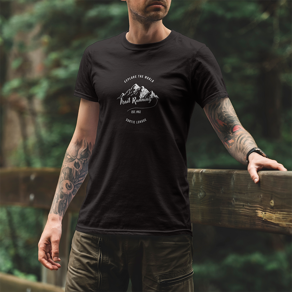 Modèle portant le t-shirt Sortie Longue noir en extérieur, vue de face, design « Explore the world » et « Trail running » avec chaîne de montagnes stylisée; tenue pour marche, sentier et déplacements.
