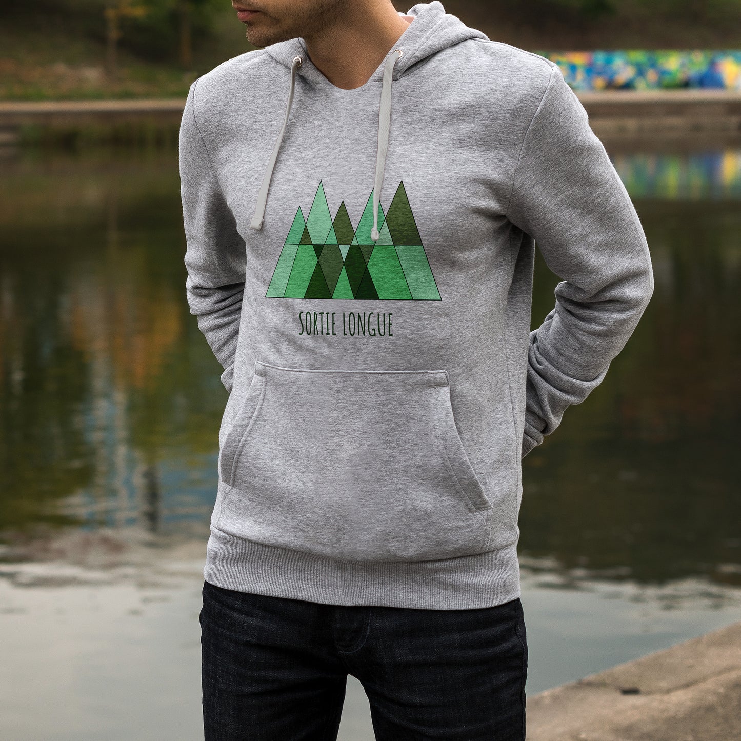 Sweat à capuche unisexe gris Sortie Longue porté en extérieur près d’un plan d’eau, logo montagne et forêt visible sur la poitrine, vêtement pour matin frais, approche et retour de terrain